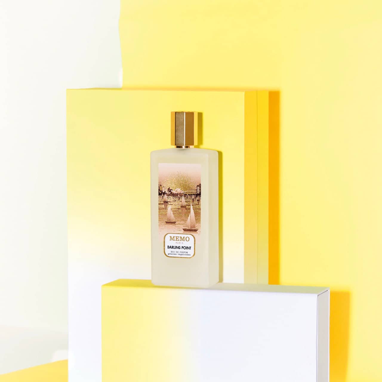 A sleek, rectangular white glass bottle of memo paris darling point eau de parfum with a gold cap and an illustrated label depicting sailboats on water.
